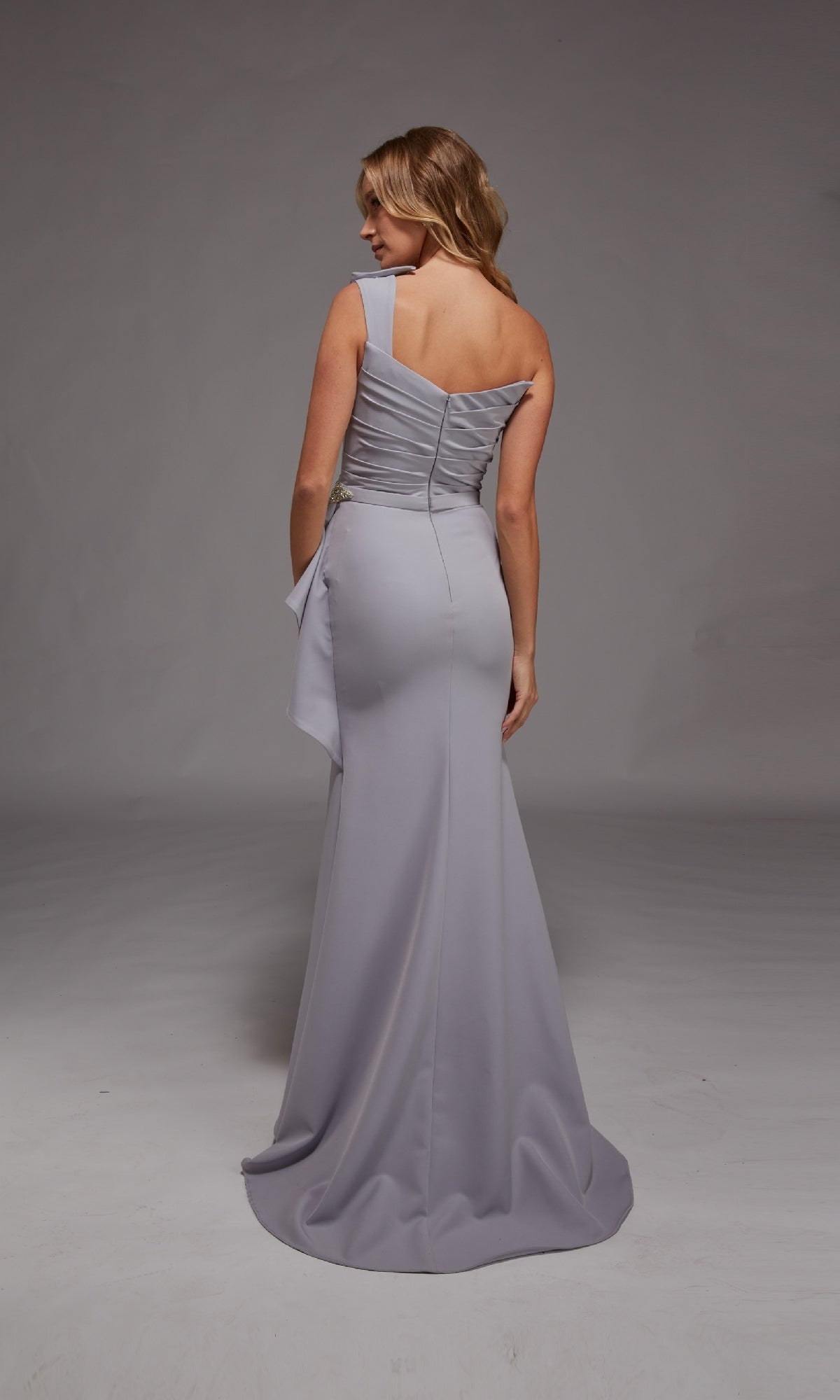 A woman stands facing away, wearing the Alyce 27812 long formal dress in light lavender, featuring a fitted waist, one-shoulder neckline, and elegant draped fabric detail.